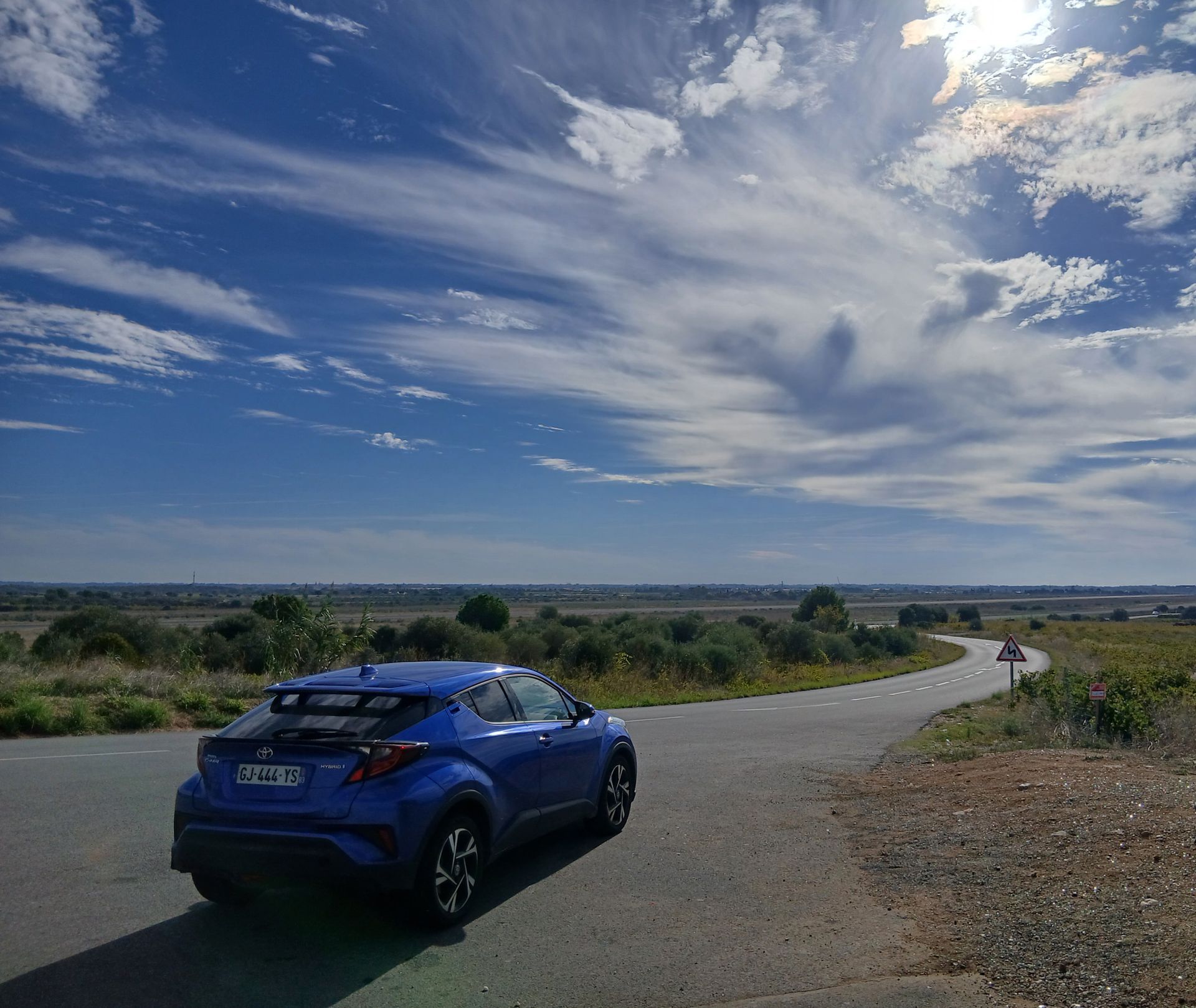 Voiture bleue sur une route sinueuse sous un ciel lumineux parsemé de nuages vaporeux.