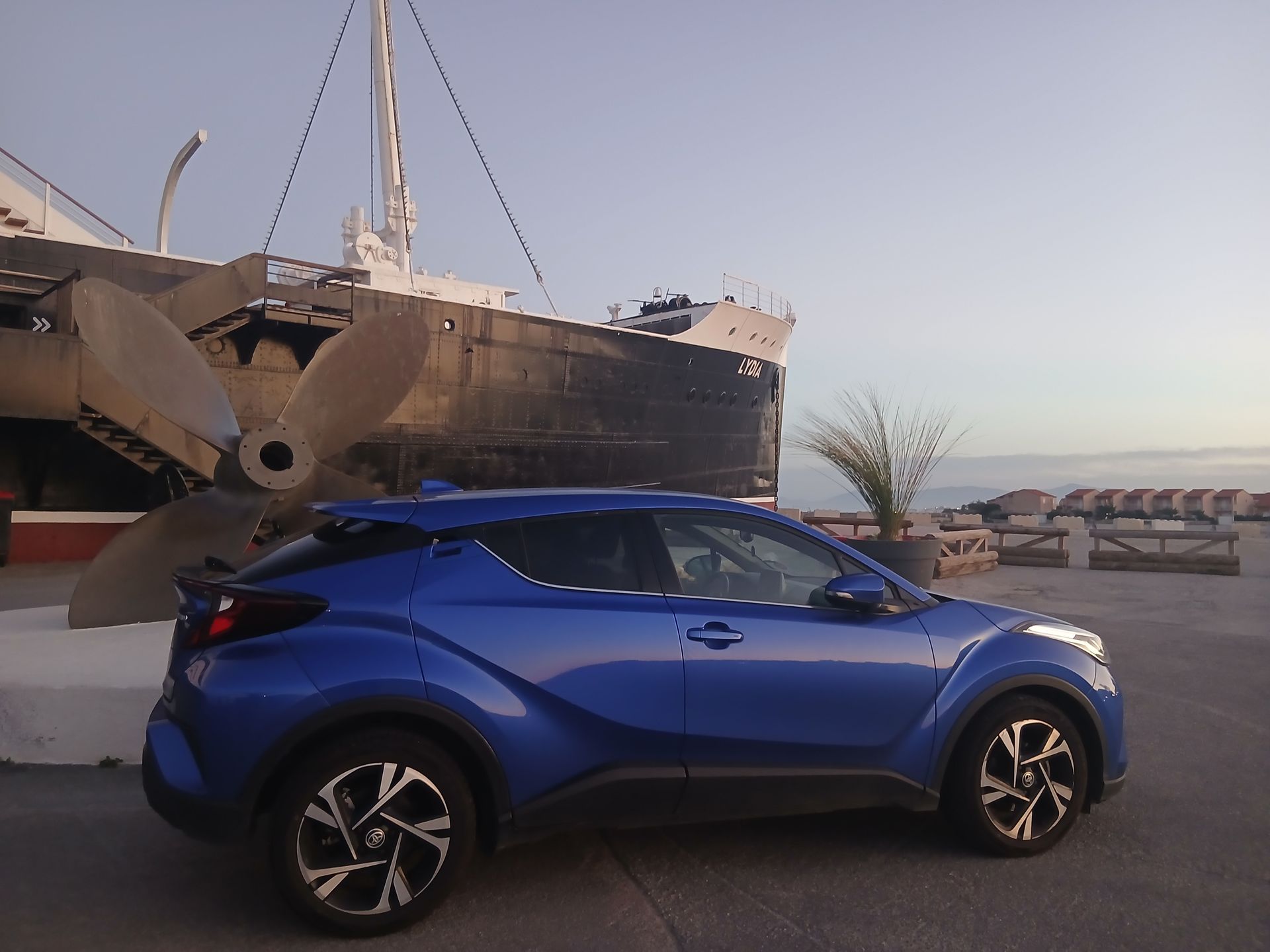 Une Toyota est garée devant un bateau à grande hélice. L'océan et des chaises de plage se dessinent à l'arrière-plan.