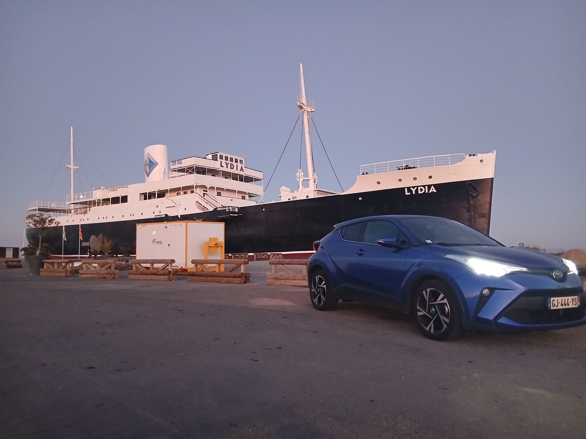 Une Toyota C-HR bleue est garée devant un grand navire au crépuscule.