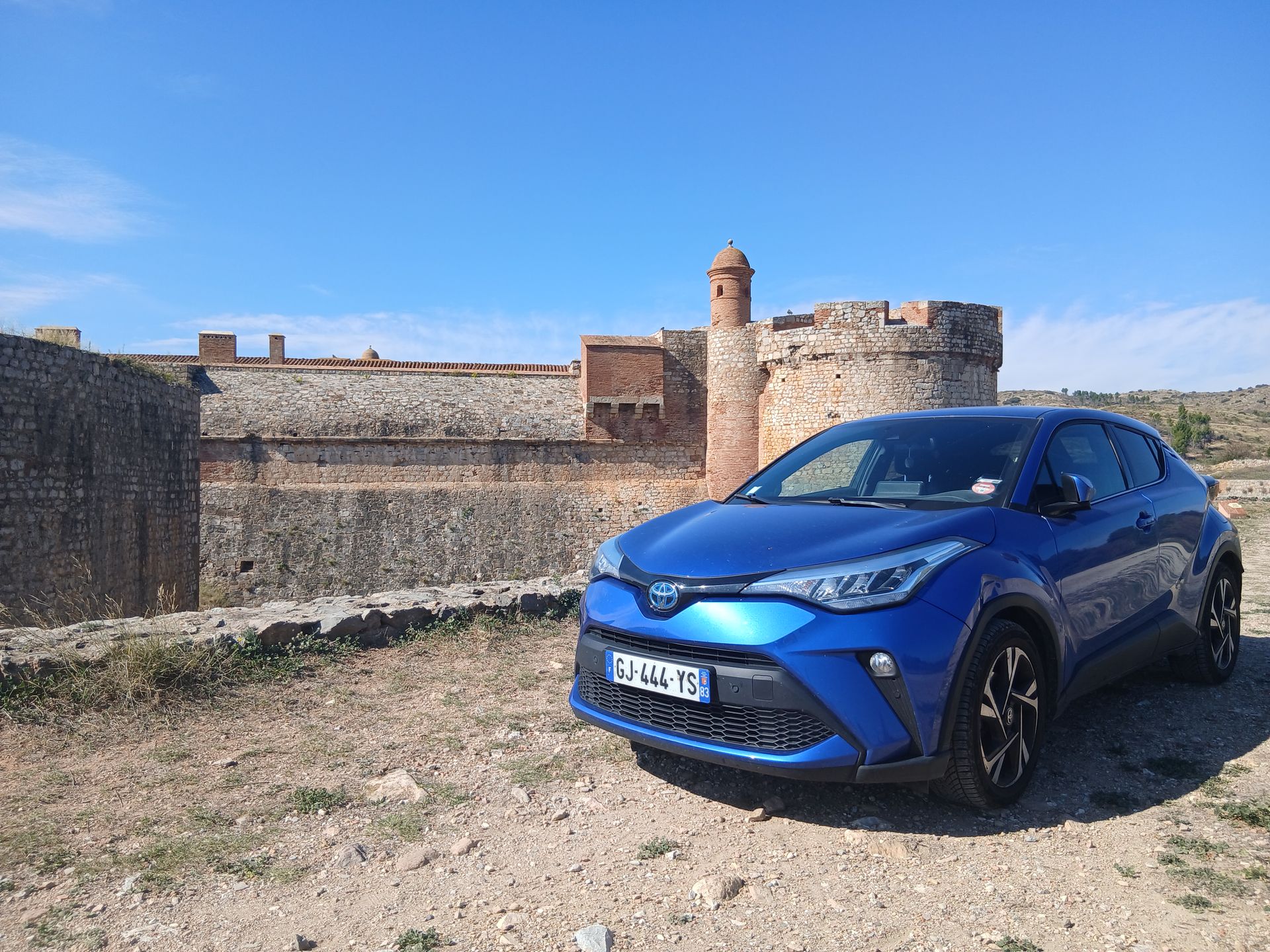 Une Toyota C-HR bleue est garée devant une forteresse en pierre sous un ciel d'un bleu limpide.