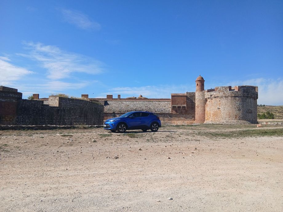 Voiture bleue garée devant un fort en pierre sous un ciel bleu limpide.
