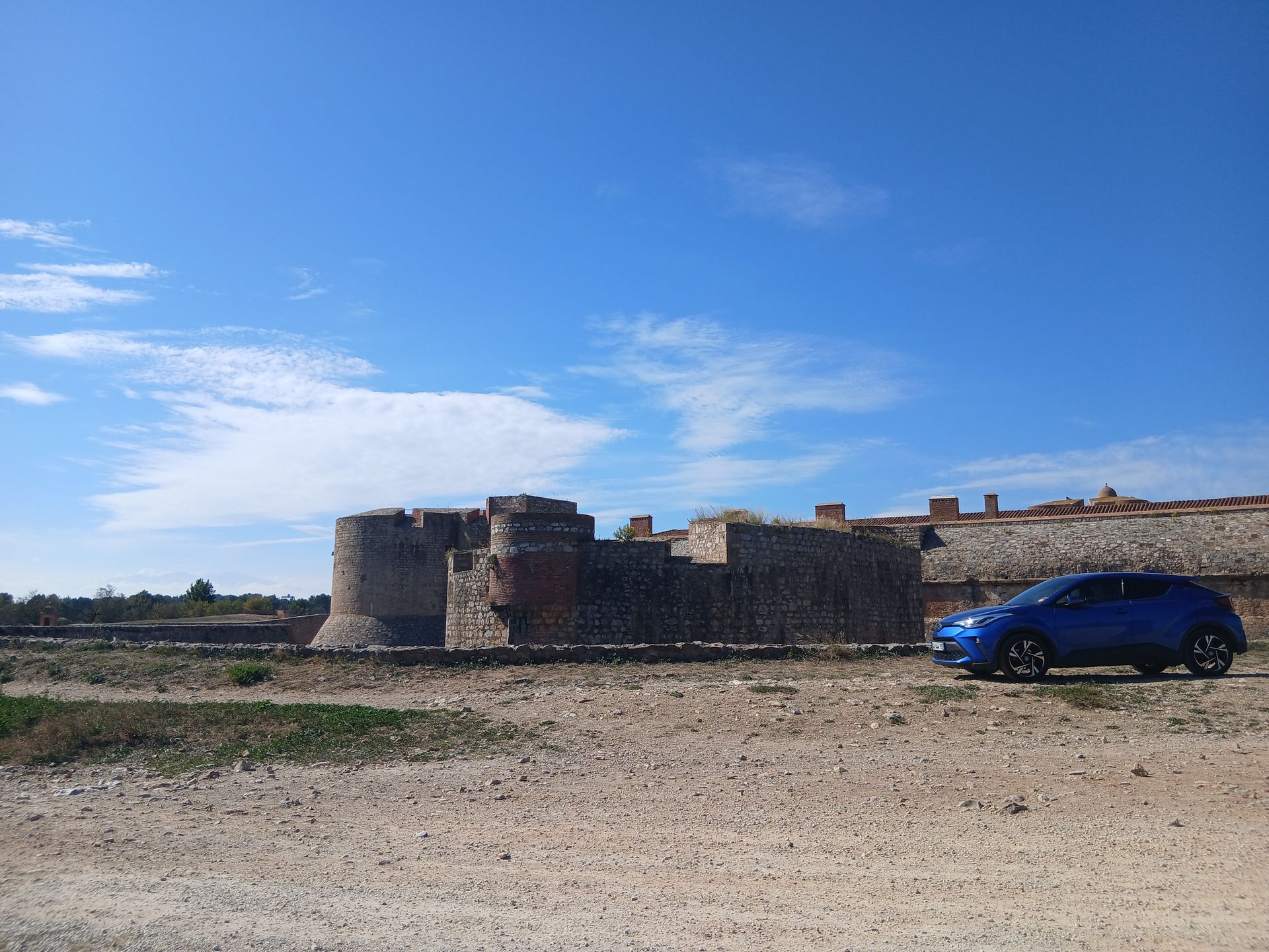 Voiture bleue garée près d'une forteresse de pierre patinée par le temps, sous un ciel d'un bleu éclatant.