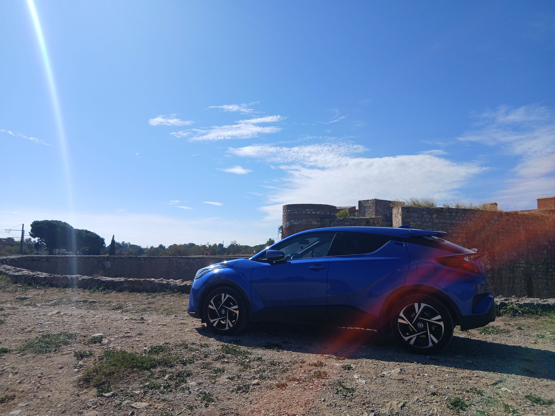 Une Toyota est garée sur un chemin de terre, avec des ruines de pierre en arrière-plan, sous un ciel bleu ensoleillé.