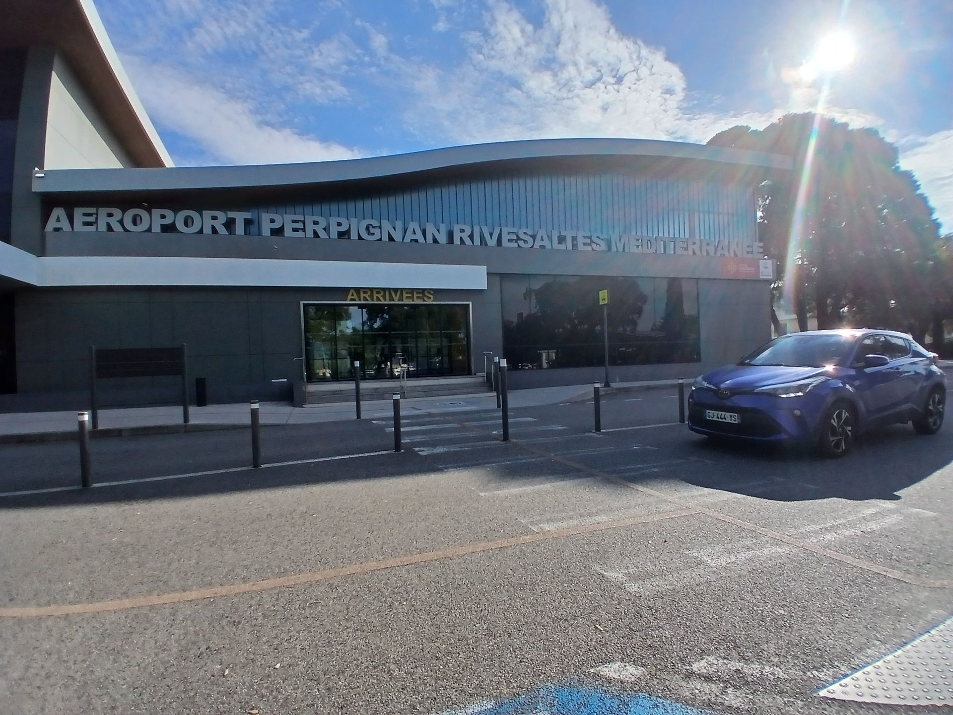 Vue extérieure de l'aéroport de Perpignan Rivesaltes avec une voiture bleue en circulation. Journée ensoleillée.