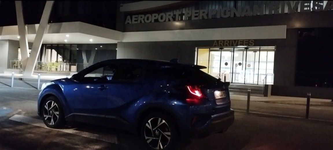 Une voiture bleue est garée devant l'aéroport de Perpignan. L'entrée est éclairée. Il fait nuit.