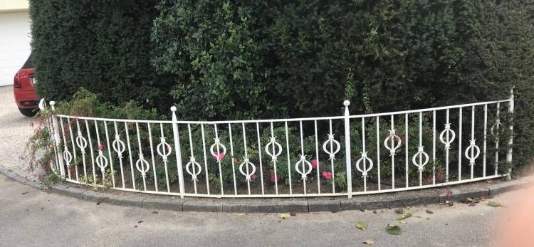 Ein weißer Metallzaun schmiegt sich an einer Straßenecke um eine Hecke; links ist ein rotes Auto teilweise zu sehen.