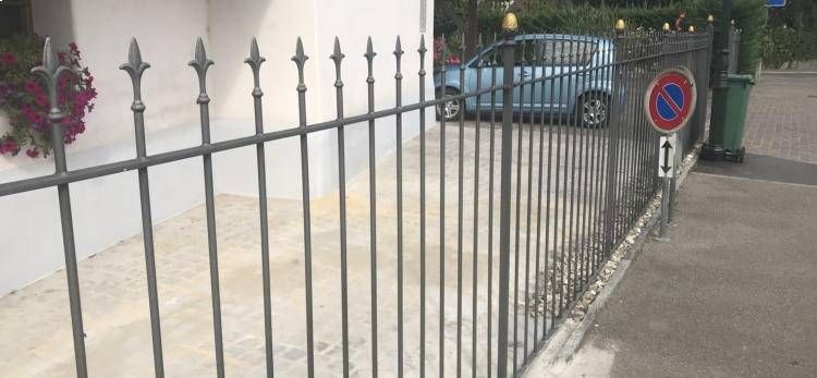 Ein Metallzaun neben einem Gehweg, im Hintergrund ein Parkverbotsschild und ein geparktes Auto.