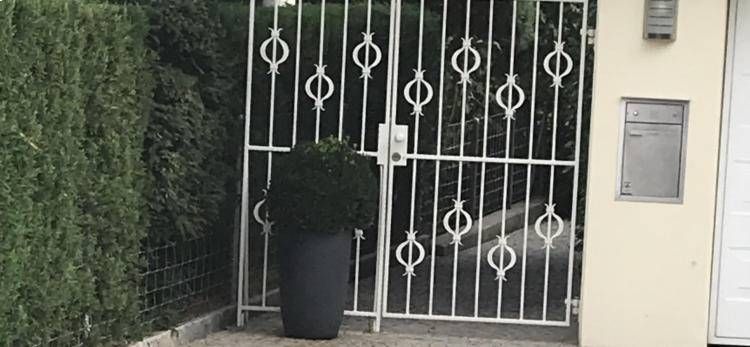 Weißes Metalltor neben einem beigen Gebäude, daneben ein Topfstrauch und ein an der Wand befestigtes Schild.