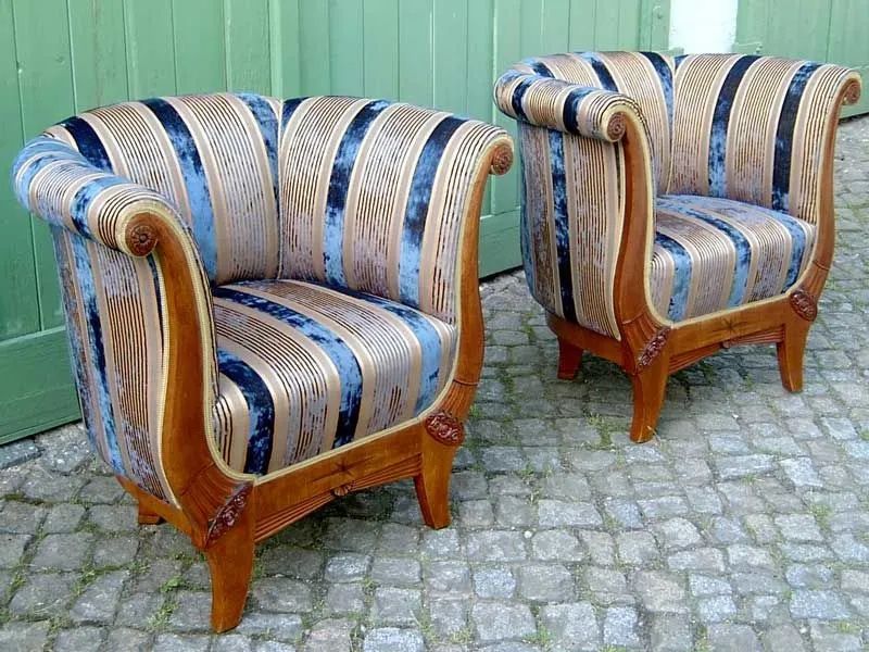 Zwei blau-cremefarbene Polstersessel mit Holzgestell auf einer gepflasterten Terrasse vor einem grünen Zaun