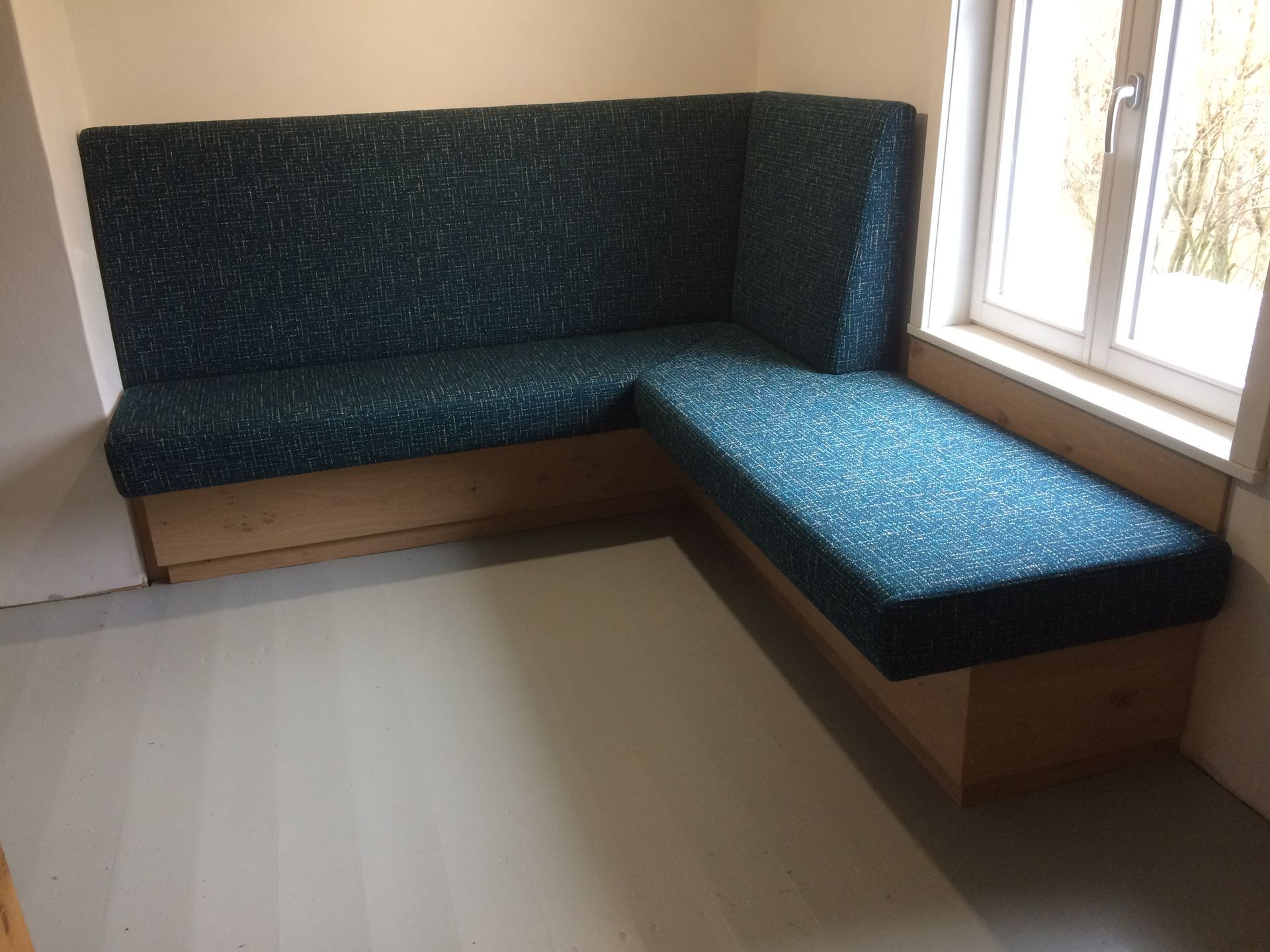 Eine L-förmige, türkis gepolsterte Sitzbank steht an einem Fenster in einem hellen Eckzimmer.