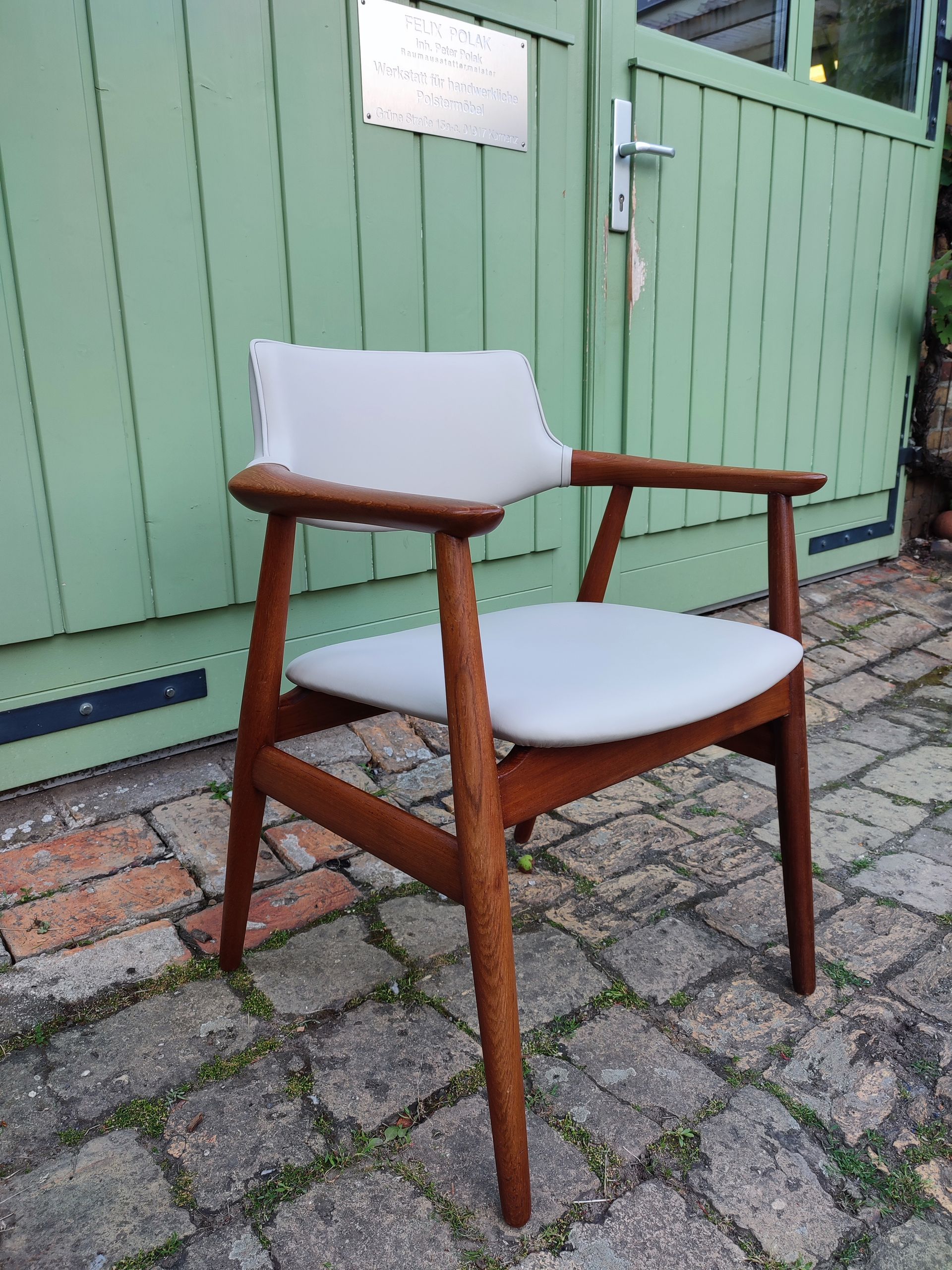 Ein Holzstuhl mit weißen Kissen auf einer gepflasterten Terrasse vor einem grünen Zaun.