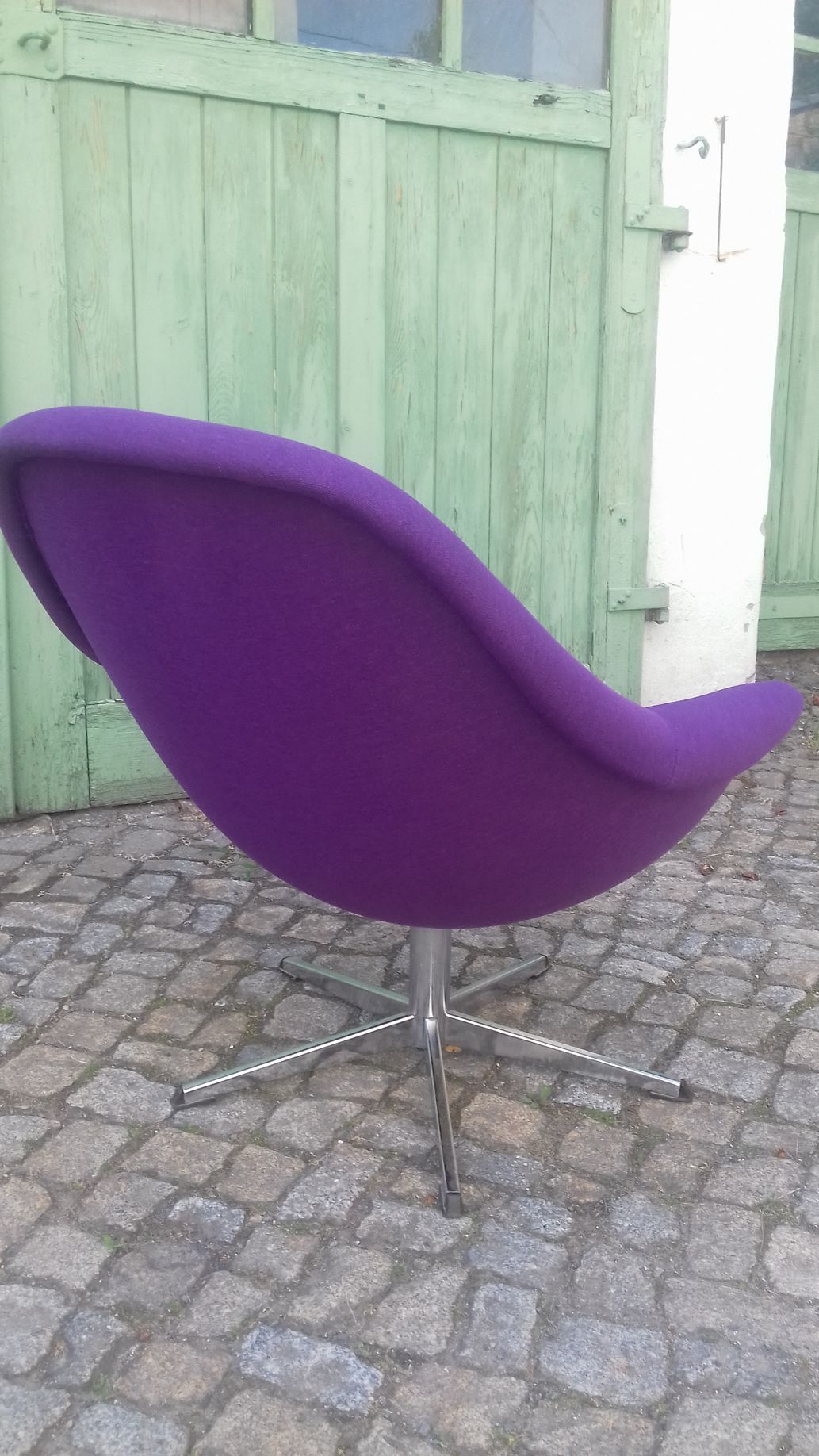 Ein moderner, lila Stuhl auf einer gepflasterten Terrasse vor einem grünen Holztor.