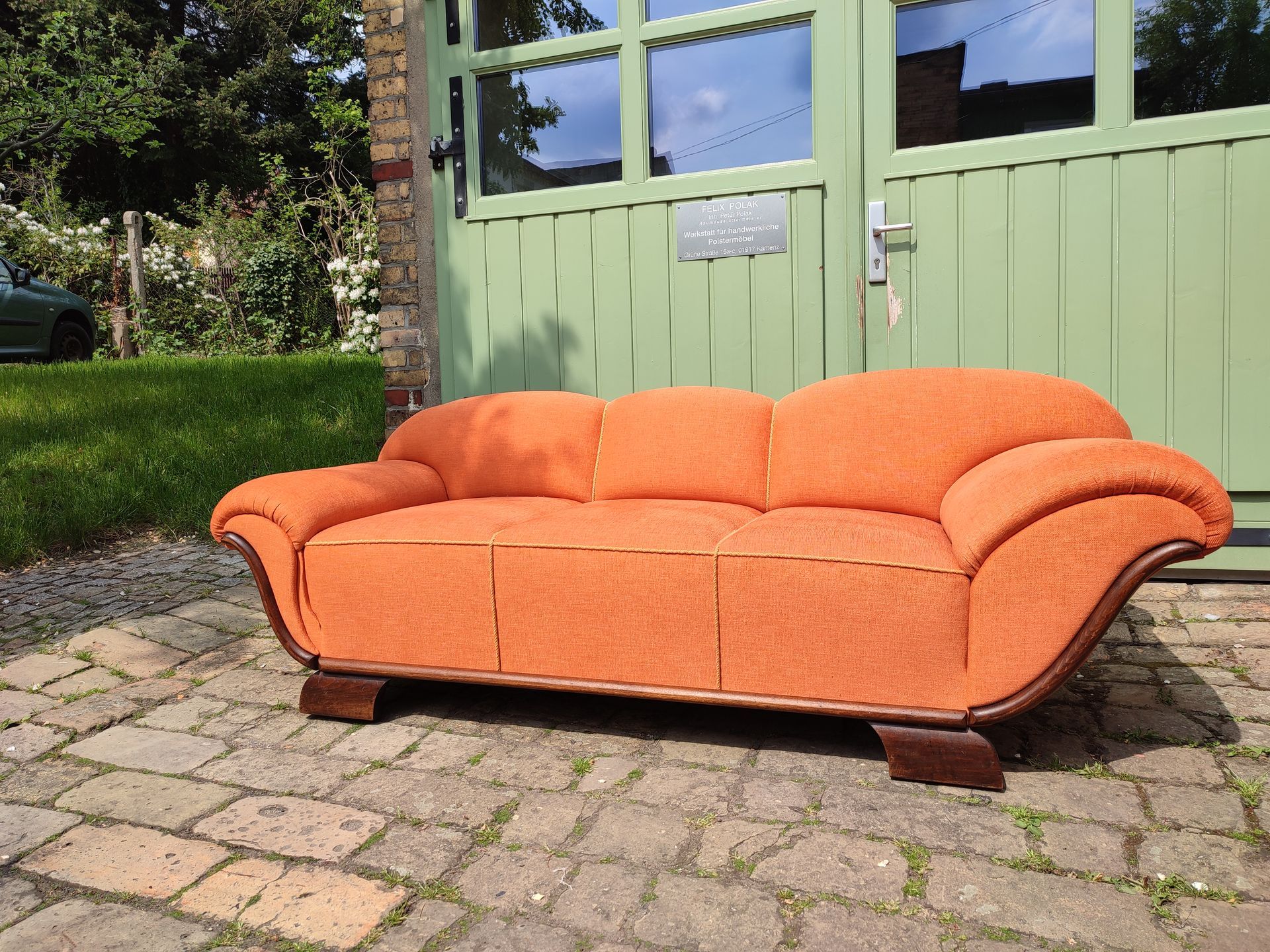 Ein orangefarbenes Sofa auf einer gepflasterten Terrasse vor einem grünen Schuppen