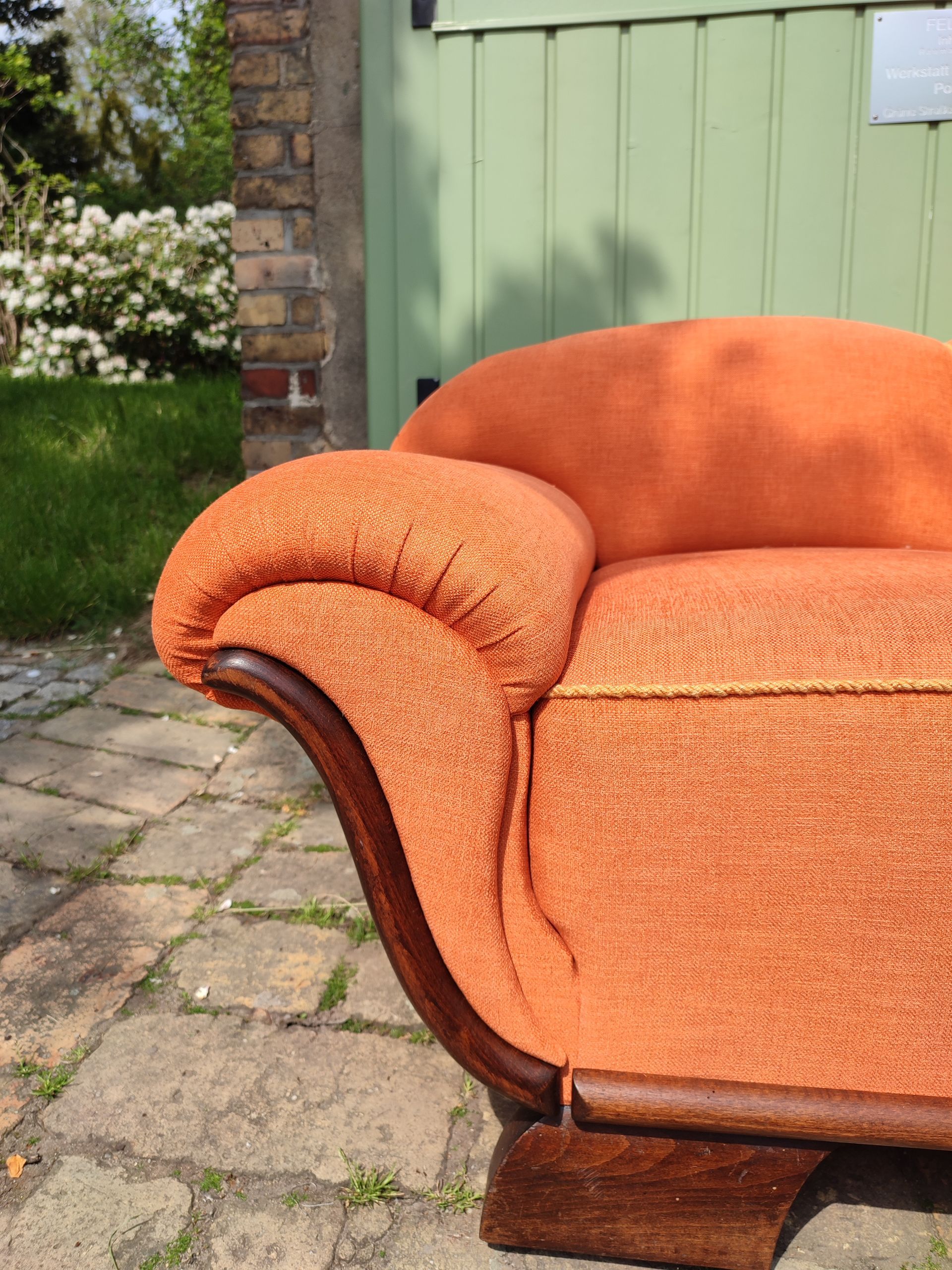 Ein orangefarbenes Polstersofa mit Holzarmlehnen steht auf einer Ziegelterrasse in der Nähe eines grünen Zauns und Blumen.