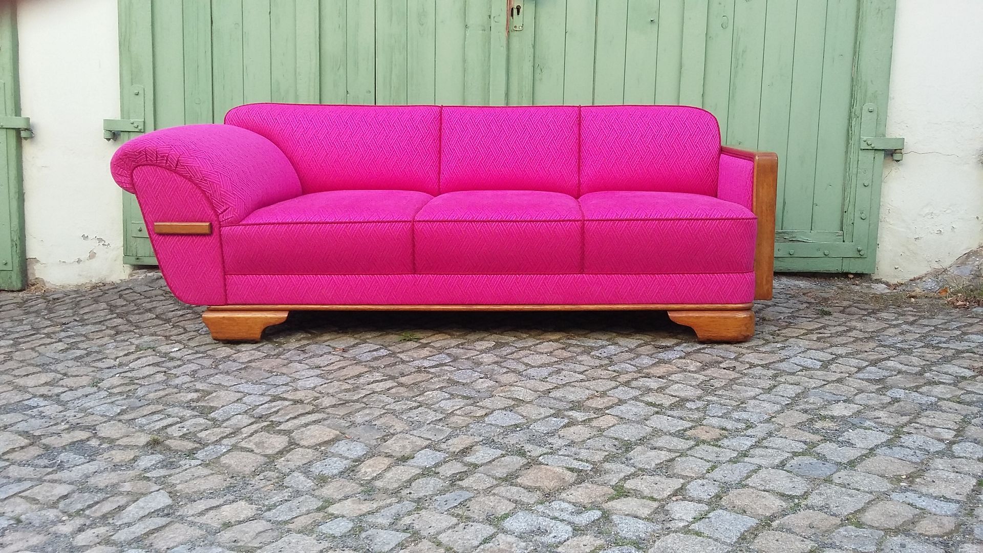 Ein leuchtend pinkfarbenes Sofa vor einer hellgrünen Wand auf einem Kopfsteinpflaster im Freien