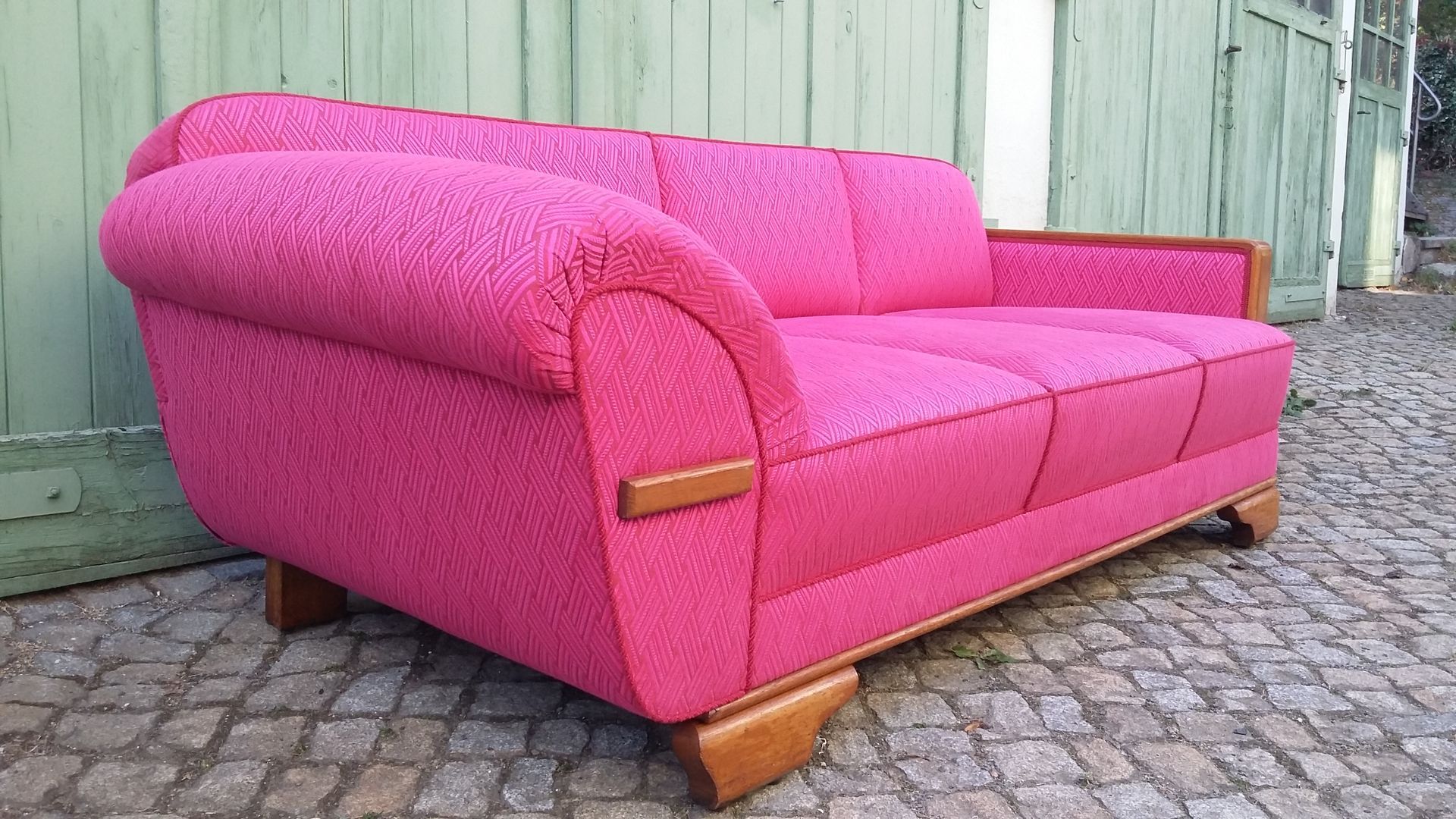 Ein leuchtend pinkfarbenes Polstersofa auf Kopfsteinpflaster neben einer hellgrünen Holzwand.