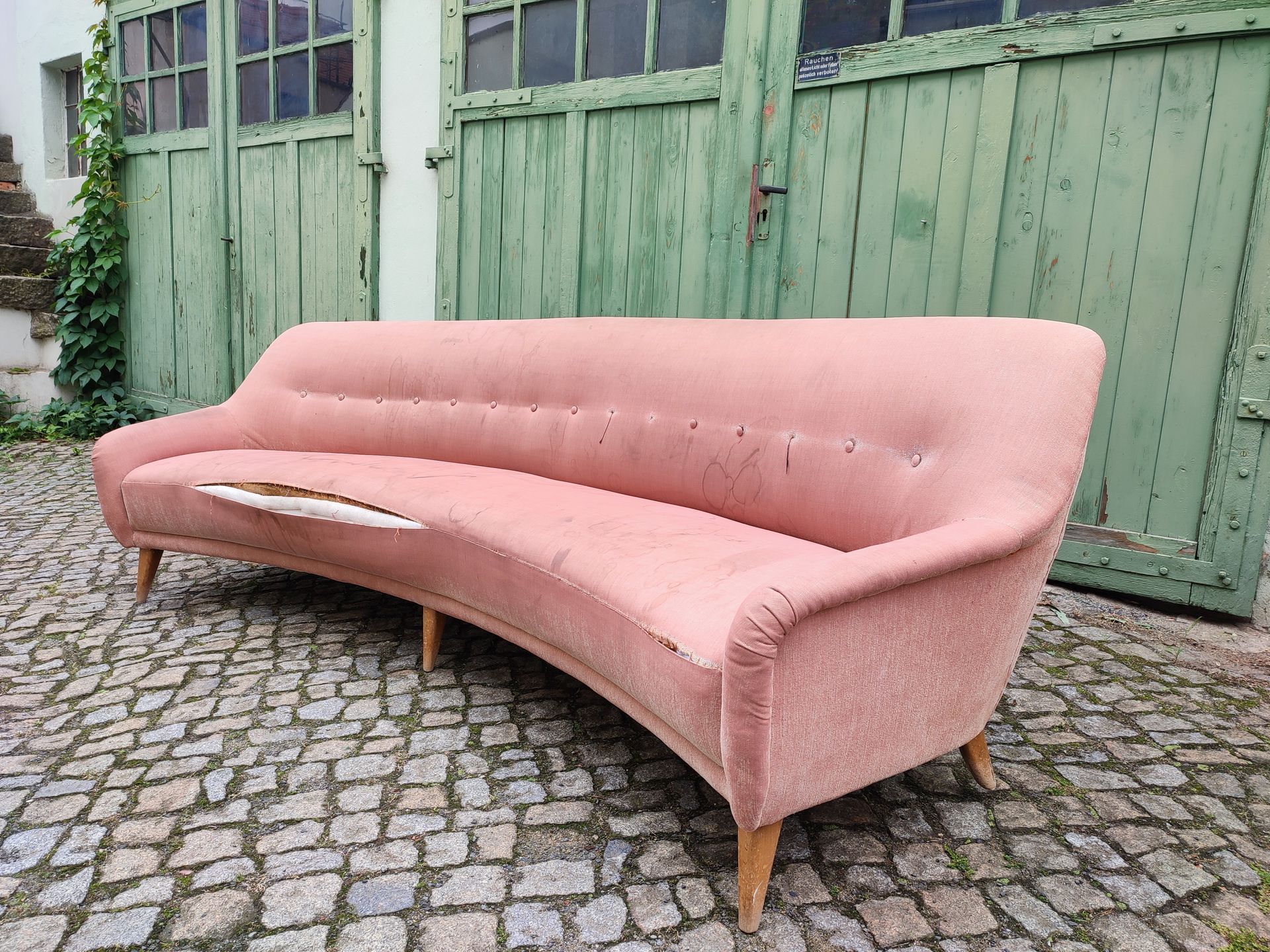 Rosa geschwungenes Sofa auf Kopfsteinpflaster vor einem grünen Holzgebäude