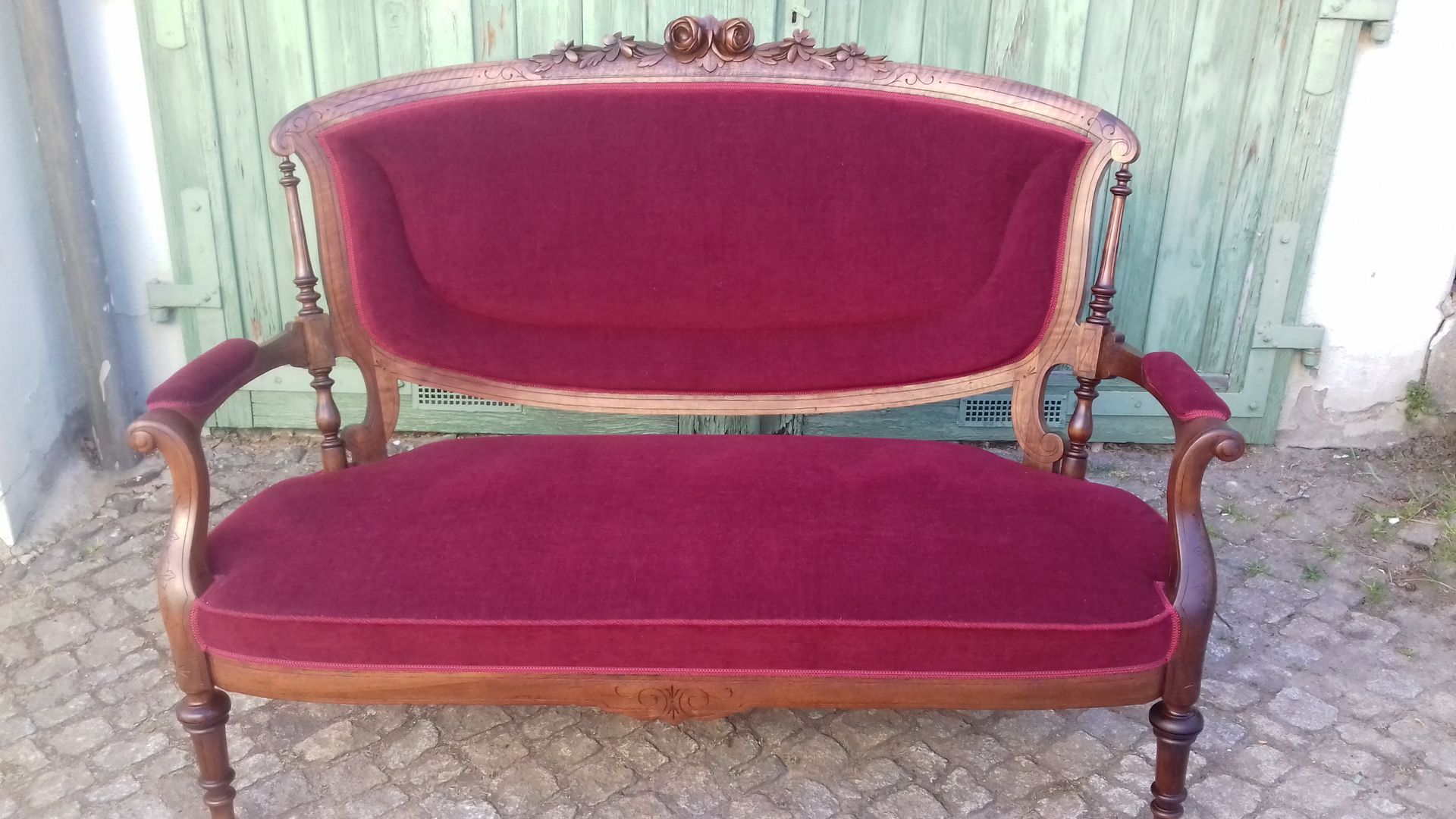 Vintage-Zweisitzer-Sofa aus bordeauxrotem Samt mit geschnitzten Holzarmlehnen und -beinen vor einer Wellblechwand