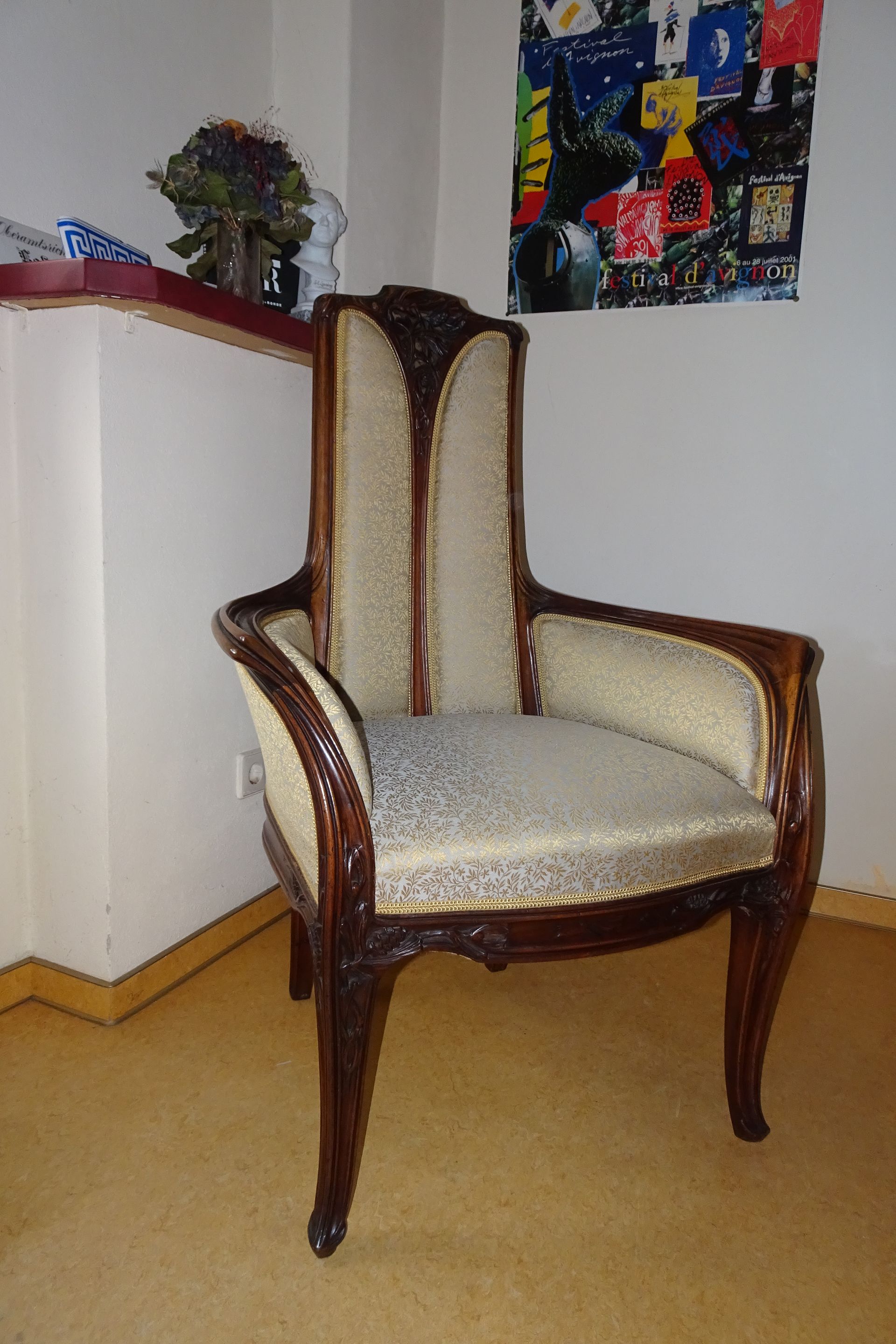 Ein antiker, cremefarbener und brauner Polstersessel steht in einer Ecke neben einem weißen Schrank und Wandbildern.