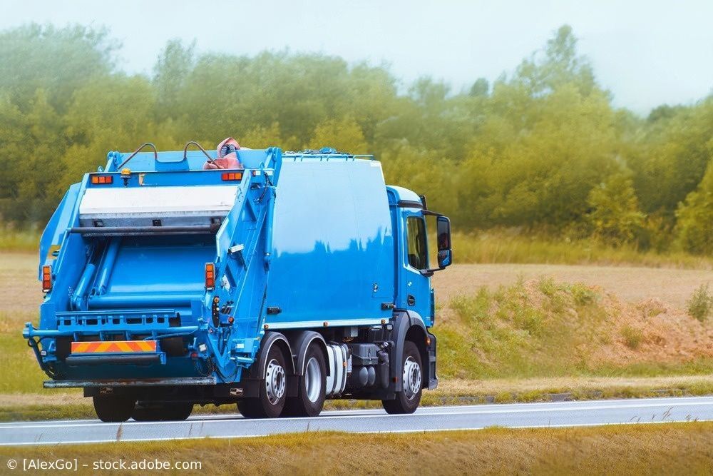 Ein blauer Müllwagen fährt auf einer Straße neben Bäumen und Gras unter einem bewölkten Himmel