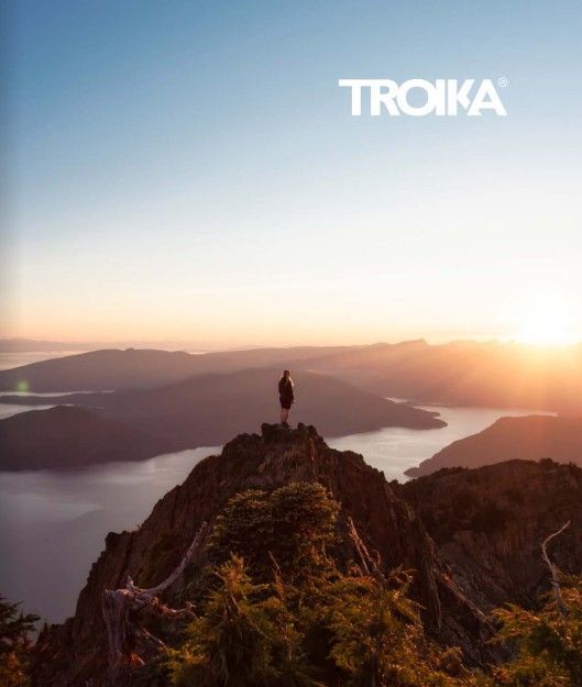 Personne au sommet d'une montagne au coucher du soleil, surplombant l'eau et les montagnes, logo « TROIKA » dans le ciel.