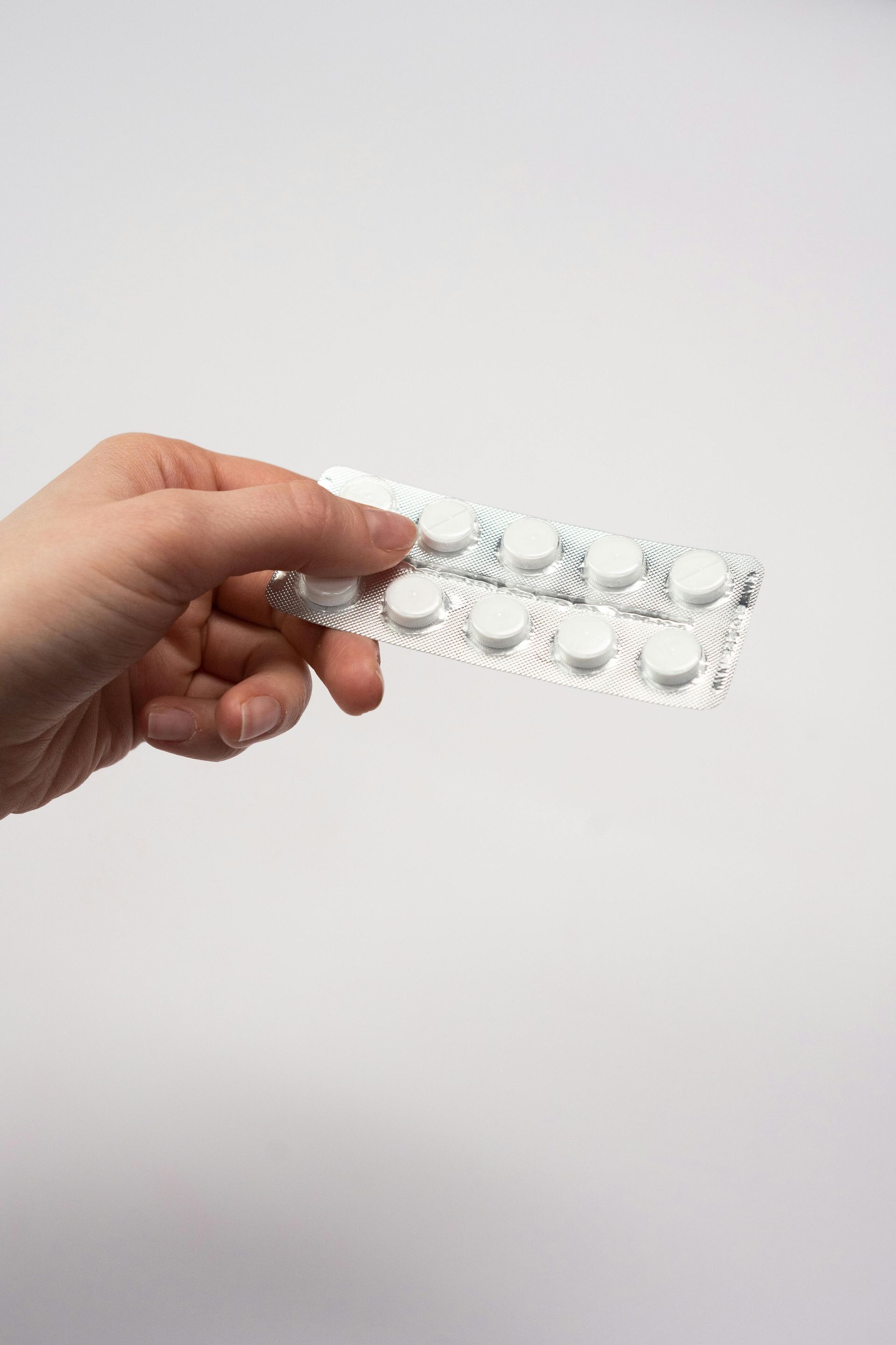 Una mano sostiene un blíster que contiene diez pastillas blancas y redondas sobre un fondo blanco liso.