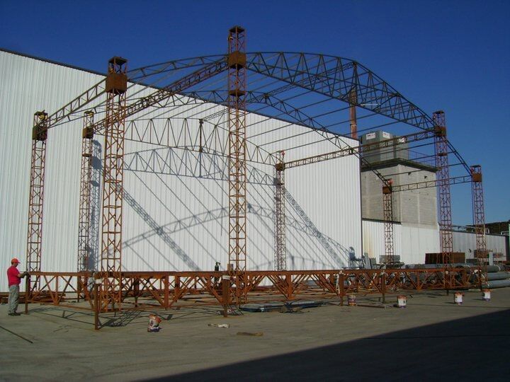 Estrutura de aço laranja sendo construída em frente a um prédio industrial branco sob um céu azul.