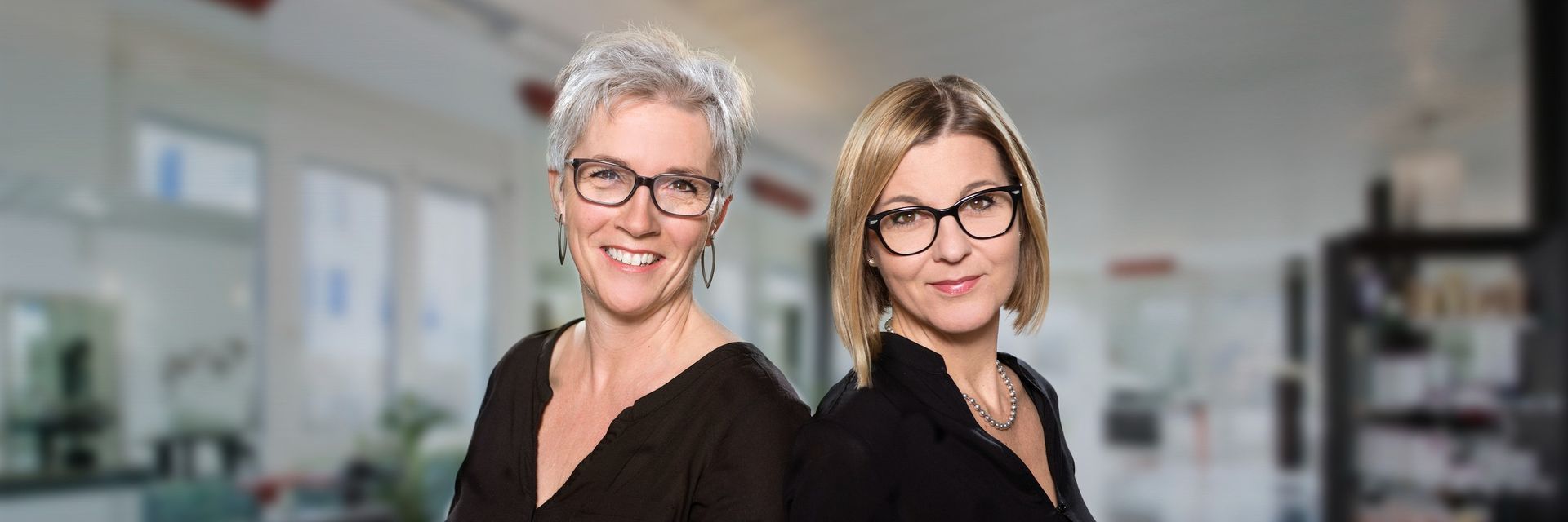 Zwei Frauen mit Brillen stehen nebeneinander in einem Raum.