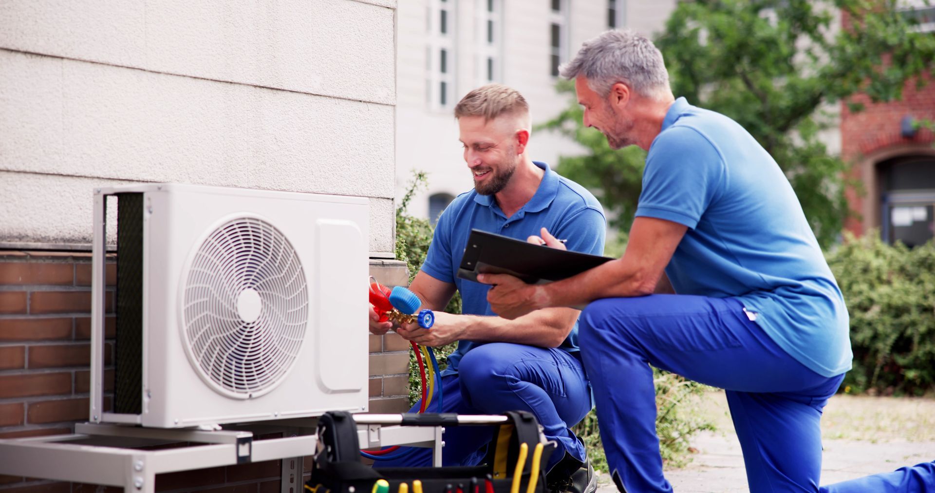 Deux techniciens en tenue professionnelle inspectent un climatiseur extérieur situé à côté d'une maison.