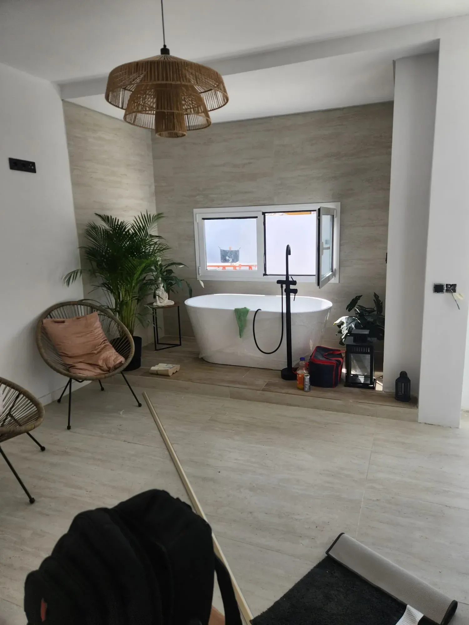 Baño luminoso con bañera independiente, silla de ratán, plantas y ventana grande.