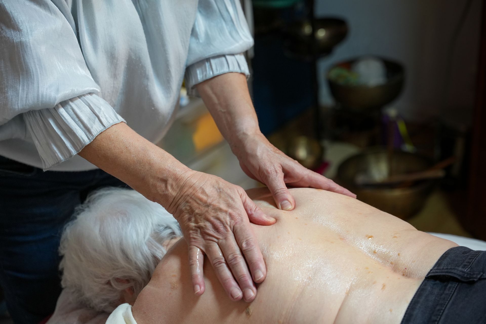 Eine Person schlägt mit einem Schlägel auf eine Klangschale, die auf dem Rücken einer liegenden Person steht. Innenaufnahme.