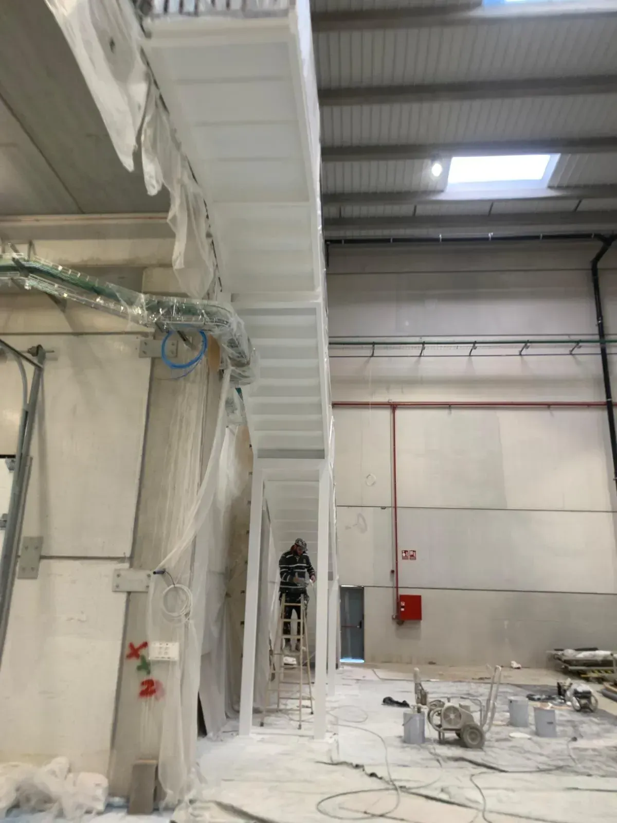 Hombre en escalera pintando vigas interiores en una gran nave industrial. Pintura blanca, techos altos.