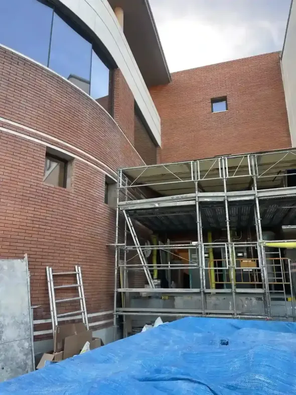 Sitio de construcción con andamios contra un edificio de ladrillo, una lona azul en primer plano y ventanas reflectantes.