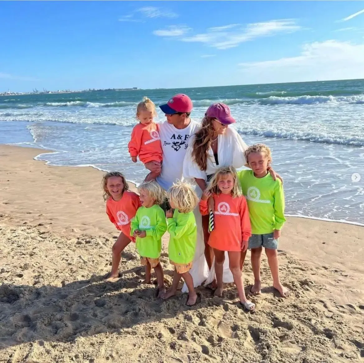 Una familia de nueve en la playa, con camisas de manga larga de colores vivos. Cielo azul y océano de fondo.
