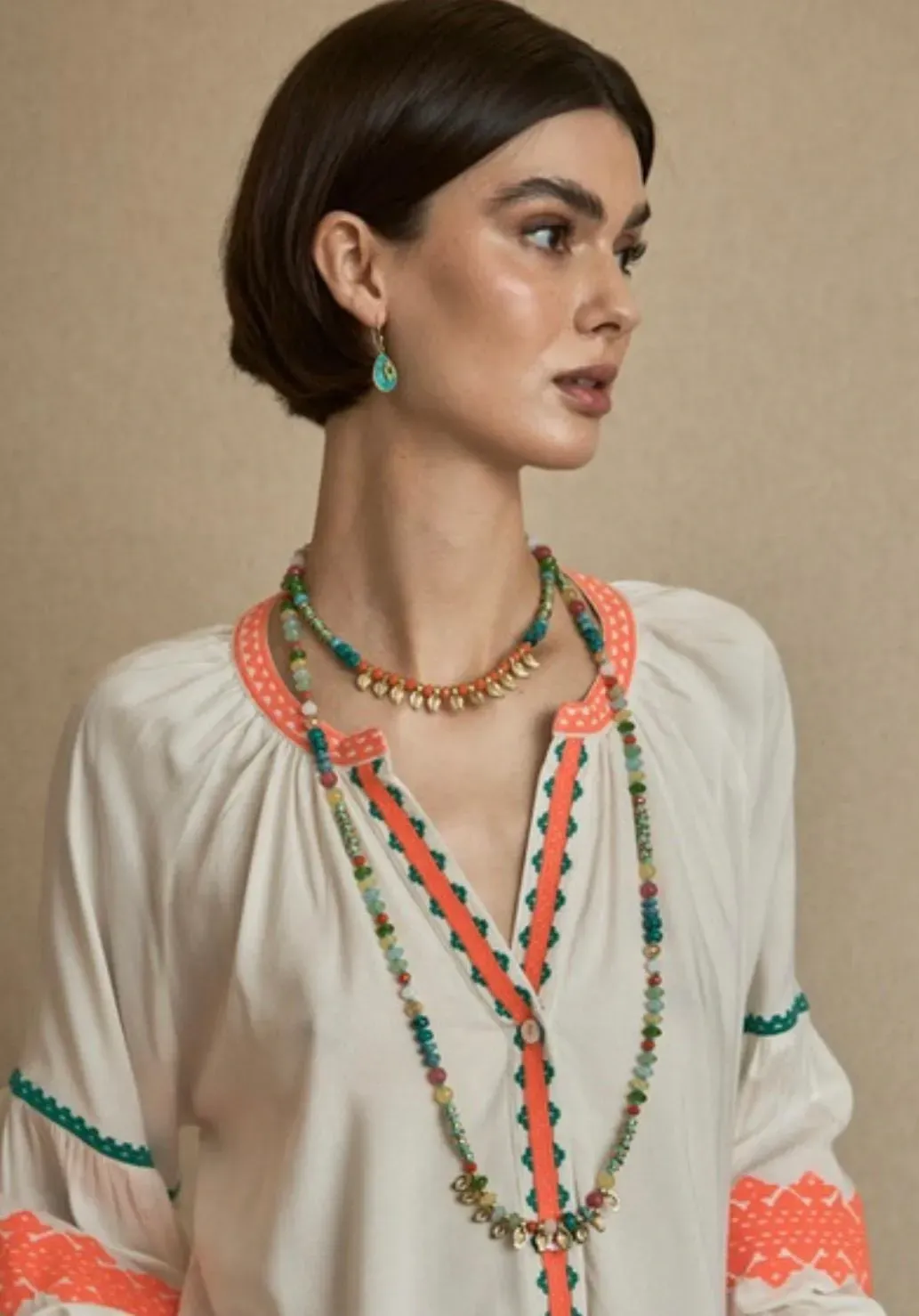 Mujer que lleva collares de cuentas de colores, aretes y una túnica blanca con ribetes naranjas y verdes.