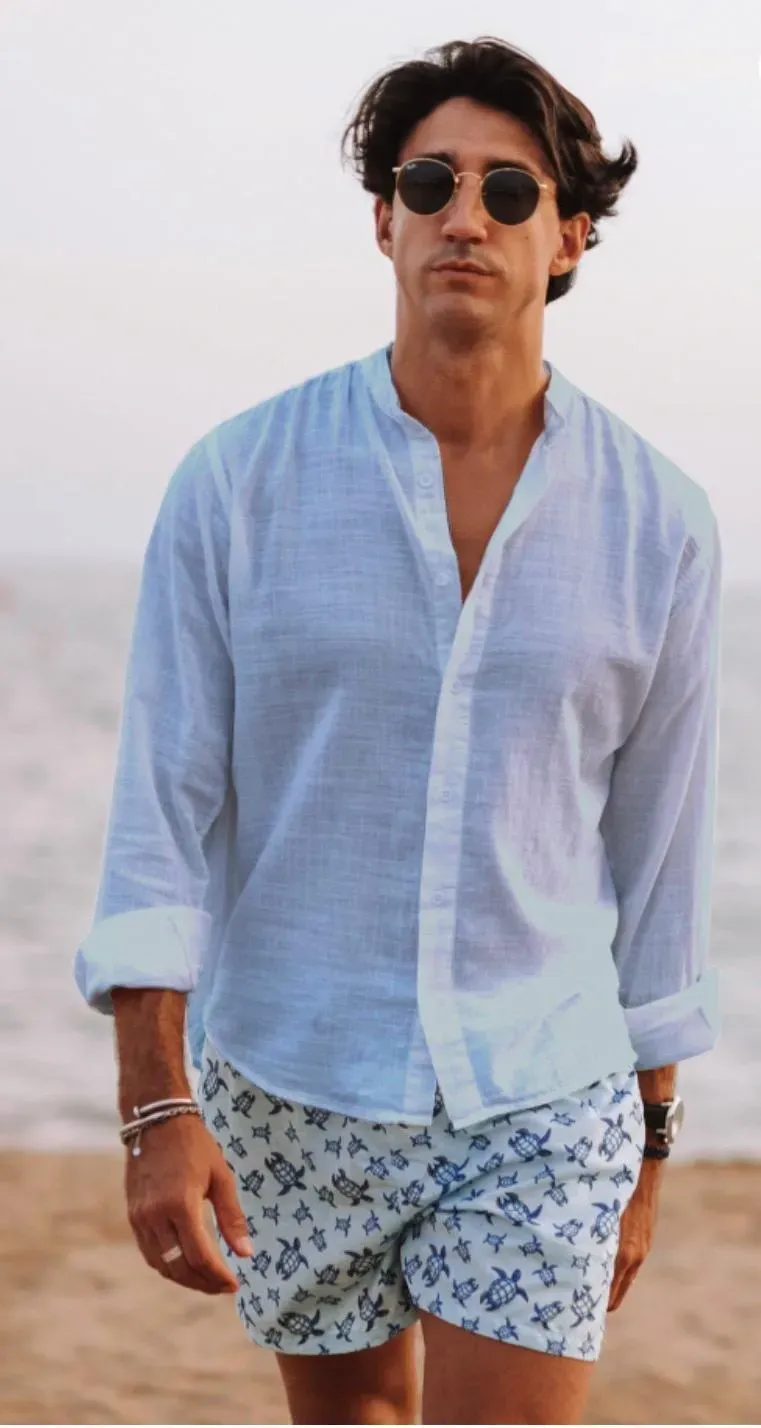 Hombre con camisa blanca y pantalones cortos estampados, de pie en la playa, con gafas de sol.