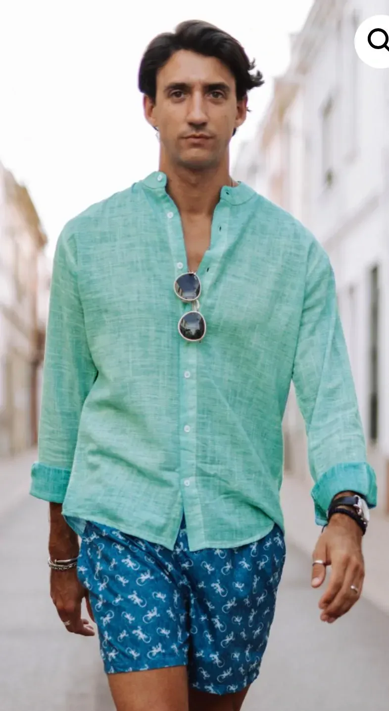 Un hombre con camisa verde azulado y pantalones cortos azules camina por una calle, con gafas de sol en la camisa.