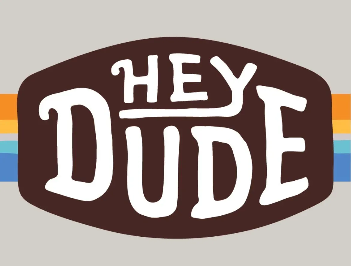 Logotipo de Hey Dude en óvalo marrón con rayas naranjas, doradas y azules sobre fondo gris.
