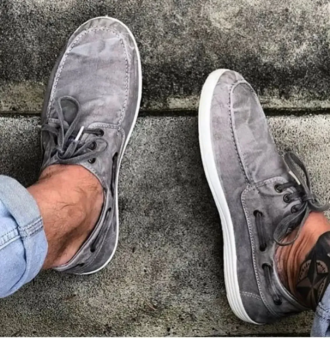 Zapatos náuticos de lona gris en los pies, apoyados sobre hormigón.
