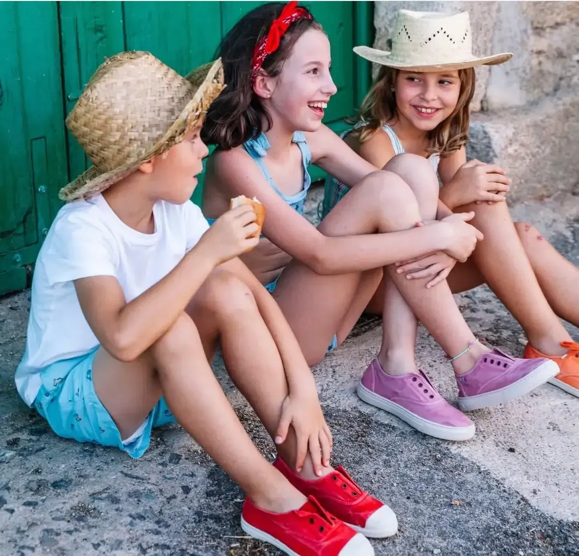 Tres niños sentados y riendo al aire libre, con sombreros de paja y zapatos coloridos.