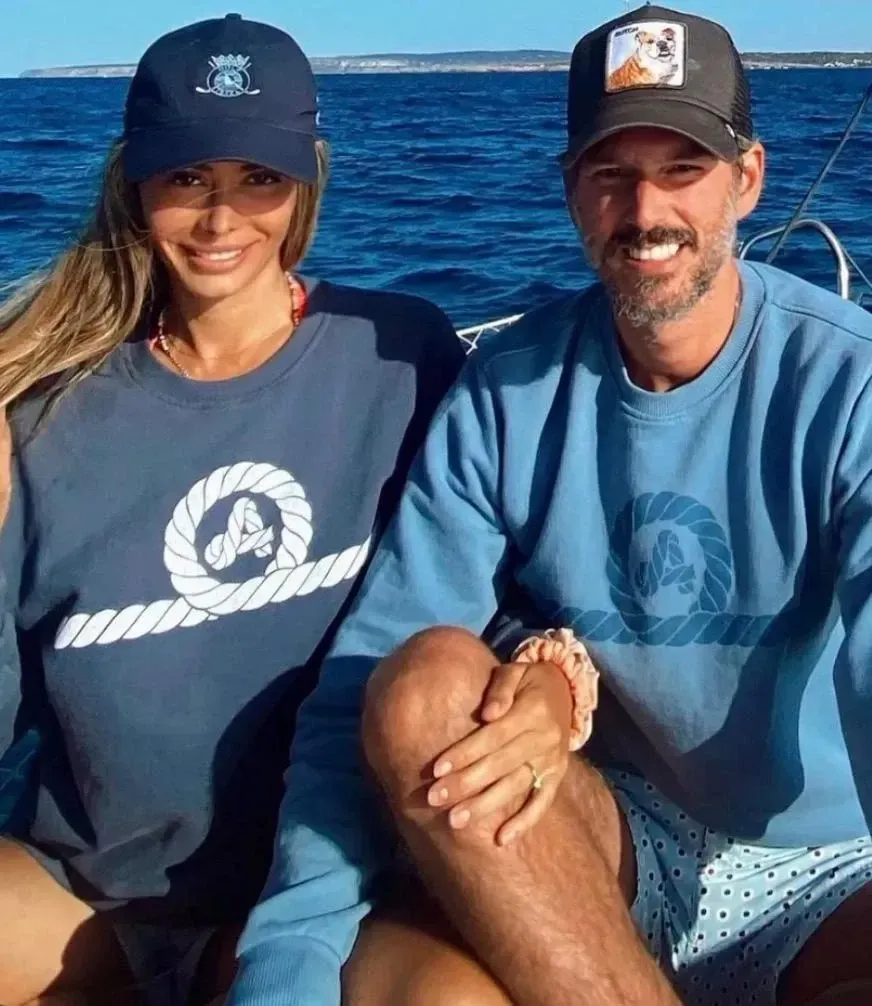 Pareja en un barco con suéteres y sombreros azules a juego, sonriendo al sol.