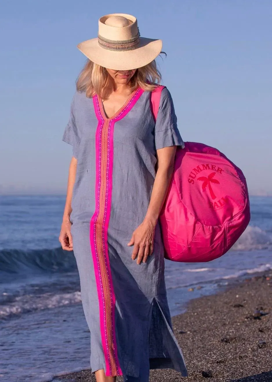 Mujer con vestido azul y sombrero, llevando un bolso rosa en una playa.