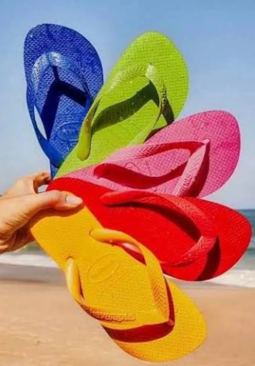 Mano sosteniendo un abanico de chanclas de colores: azul, verde, rosa, rojo y amarillo en una playa.