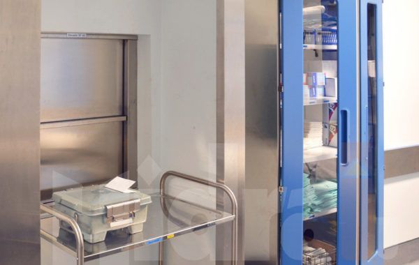 Pasillo de hospital con un carrito de acero inoxidable y una puerta con marco azul que conduce a una habitación.