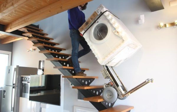Hombre subiendo una lavadora por una escalera moderna en una casa luminosa.