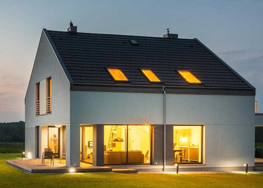 Maison moderne au crépuscule, baignée d'une lumière chaude filtrant par les fenêtres et les puits de lumière du toit.