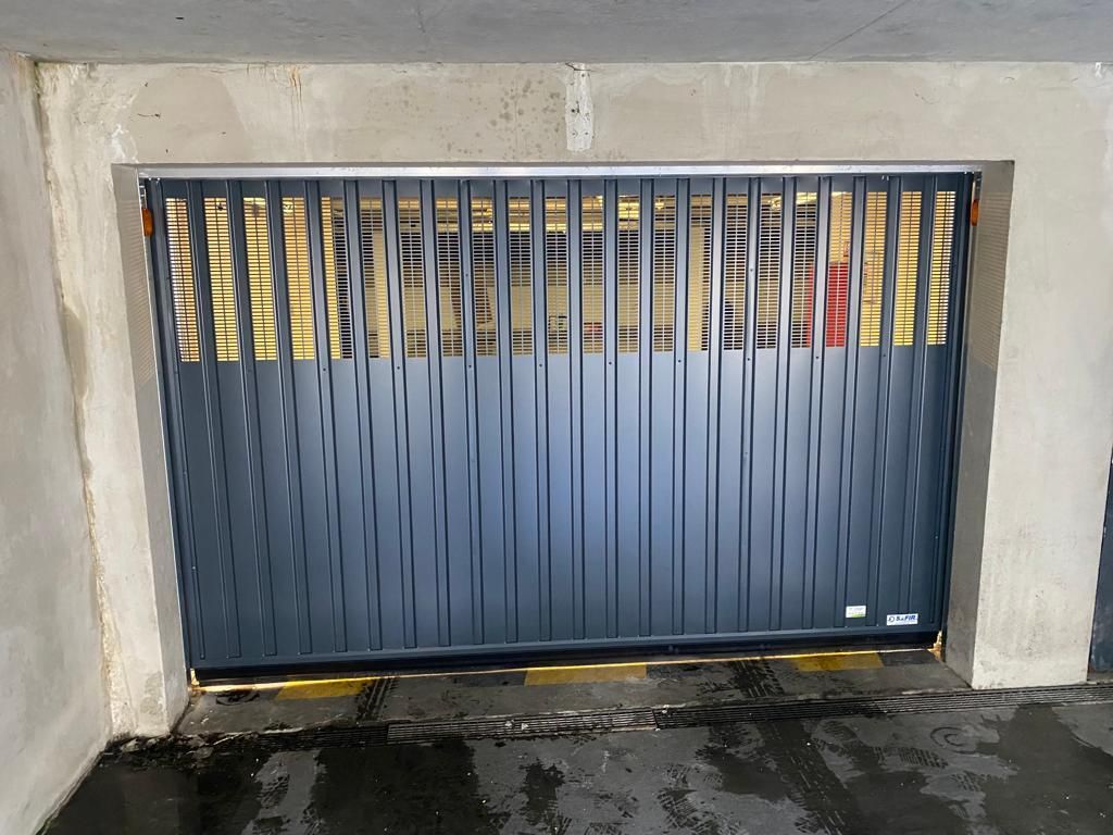 Porte de garage bleue à lames verticales encastrée dans un mur en béton, avec un trottoir mouillé devant.