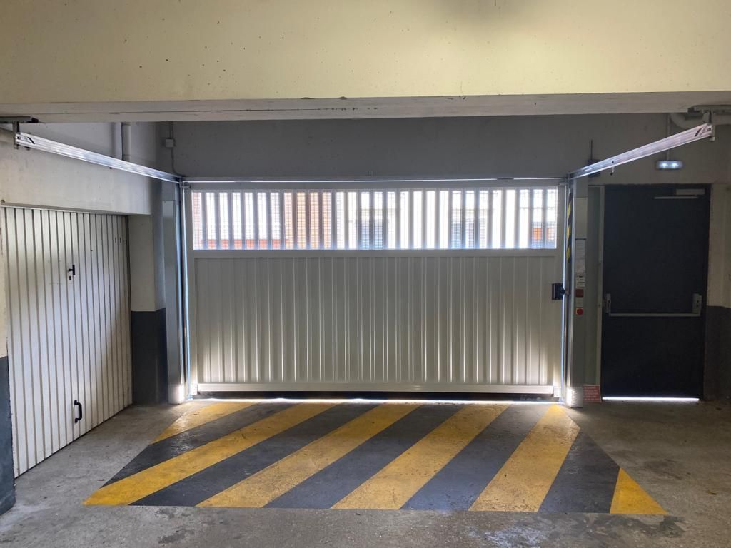 Place de parking couverte avec rampe à rayures jaunes et noires et portail blanc fermé dans un garage.