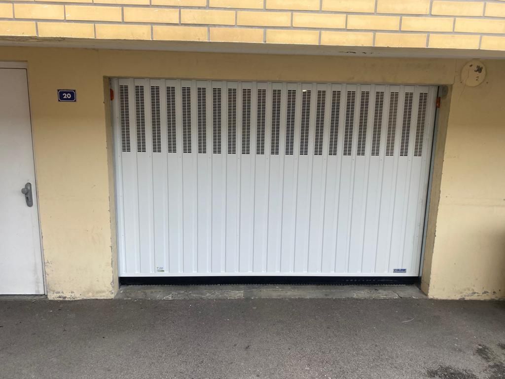 Porte de garage blanche avec petits panneaux de ventilation encastrée dans un mur de briques beiges.