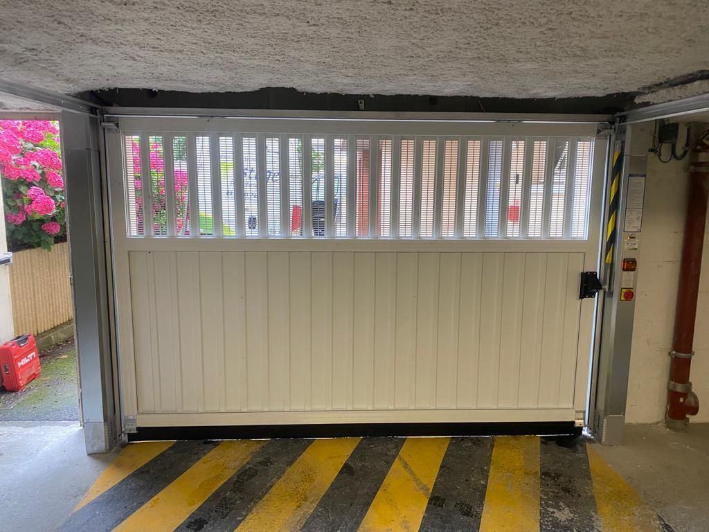 Porte de garage beige fermée avec persiennes vitrées dans une aire de stationnement, au-dessus de bandes de sol jaunes et noires.
